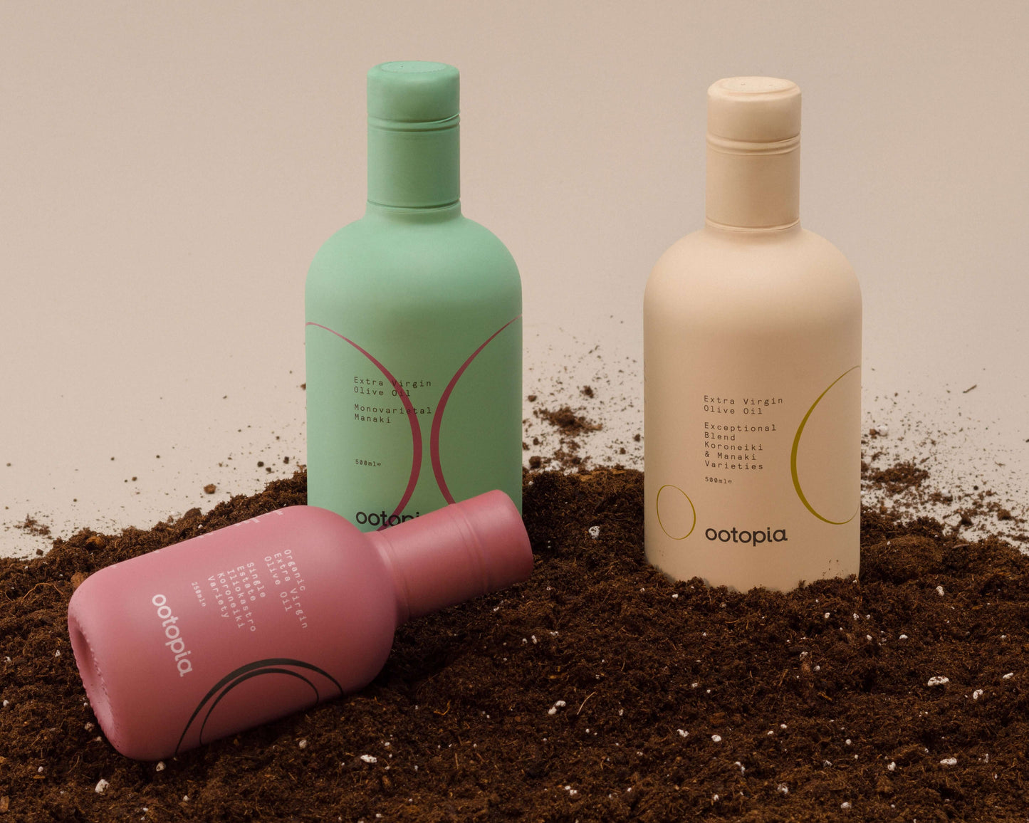 Ootopia's three olive oil bottles in pastel colors resting on soil backdrop, showcasing the Koroneiki, Manaki, and Estate Blend.